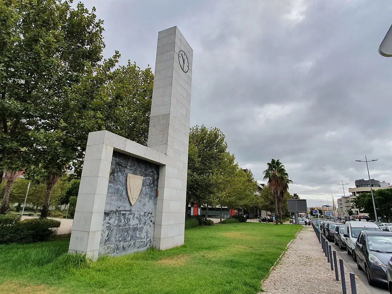 Jardim da Avenida de Moscavide, com relvado, árvores e o monumento de pedra